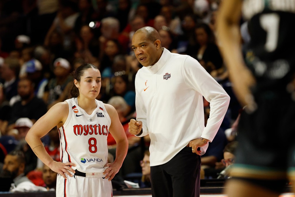 Washington Mystics head coach Sydney Johnson stands next to guard Georgia Amoore. He is leaning slightly in her direction as he talks to her, and she is standing with her hands resting on her hips.