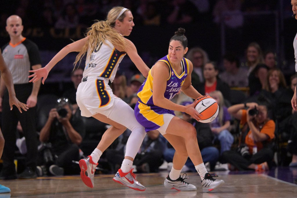 Los Angeles Sparks guard Kelsey Plum holds the ball on the perimeter with two hands and looks for a pass. Indiana Fever guard Lexie Hull defends her tightly, bumping Plum's back with her leg.