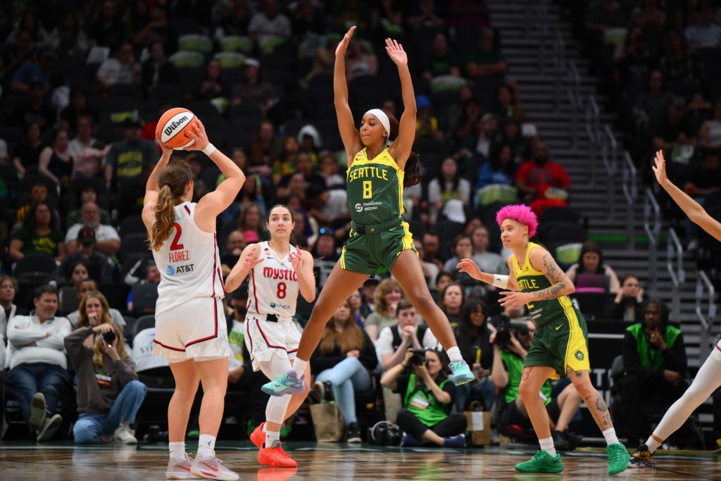 Washington Mystics point guard Alicia Flórez holds the ball with both hands over her head. Fellow point guard Georgia Amoore has her hands ready for a pass, and a Seattle Storm defender jumps in the air to dissuade a pass over the top.