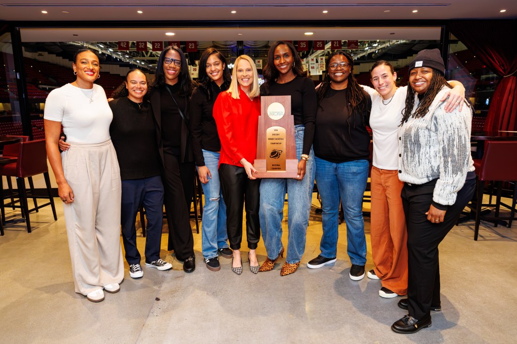 A group of nine women stand in a line, holding a trophy and smiling.