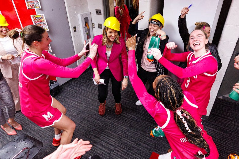 Happy players in pink uniforms surround Maryland head coach Brenda Frese who is wearing a yellow hard hat