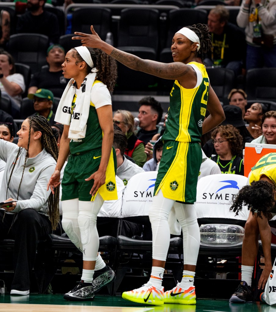 Zia Cooke holds 3-point finger guns on both sides by her hips while Jordan Horston points a 3-point finger gun with her left hand, celebrating on the bench.