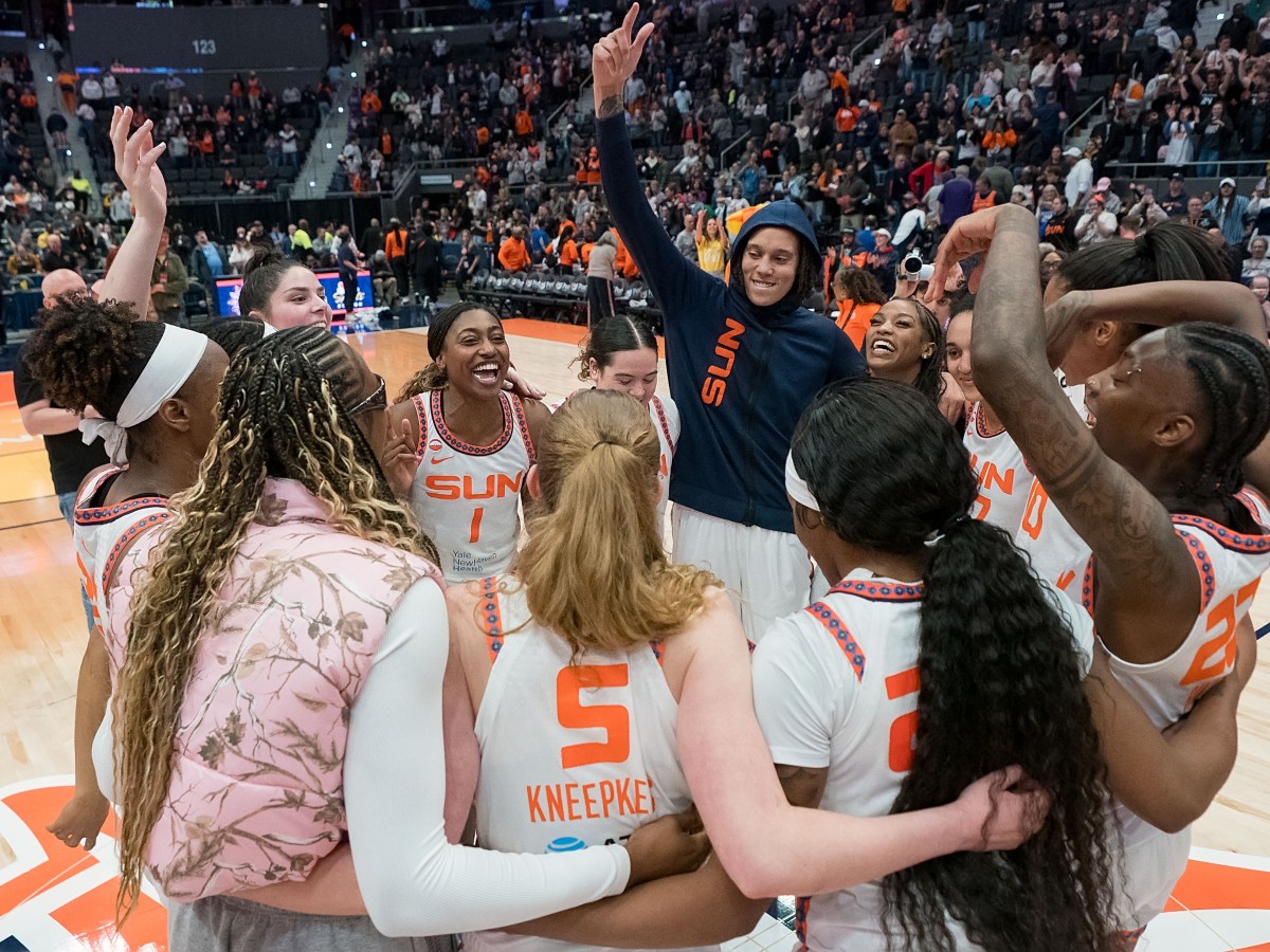 Connecticut Sun players are in a circle with smiles on their faces. Brittney Griner has her navy blue Sun hoodie on with the hood over her head, raising her hand in the air.