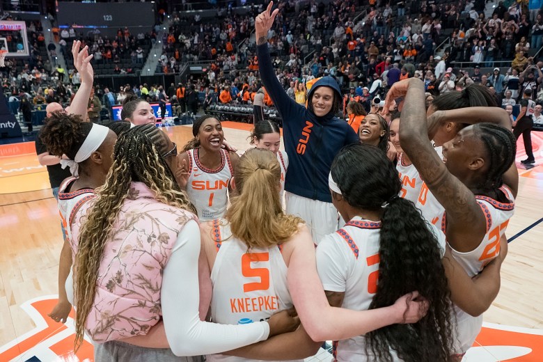 Connecticut Sun players are in a circle with smiles on their faces. Brittney Griner has her navy blue Sun hoodie on with the hood over her head, raising her hand in the air.