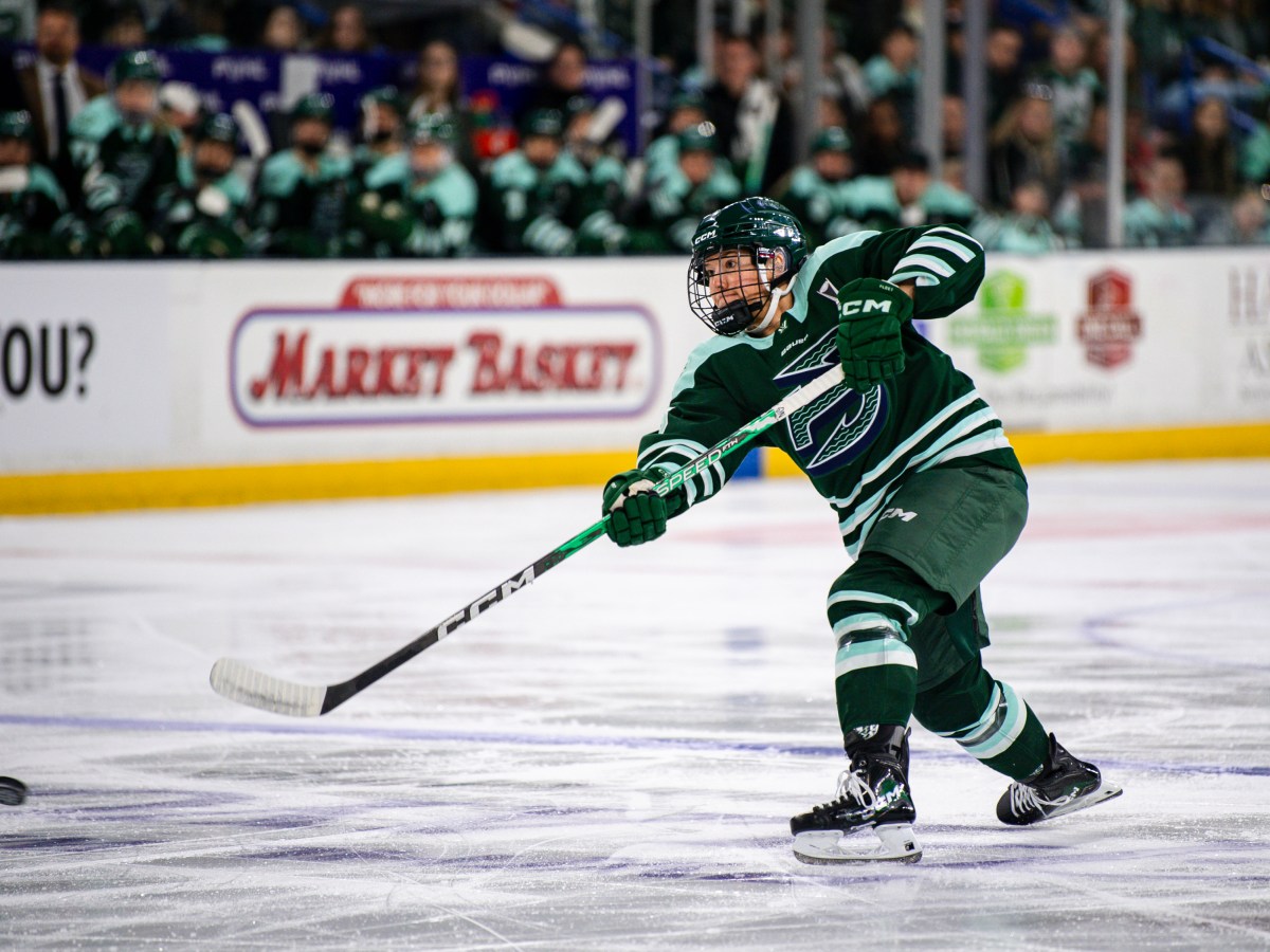 Winn leans forward and follows through with her stick as the puck sails toward the goal. She is wearing a green home uniform.