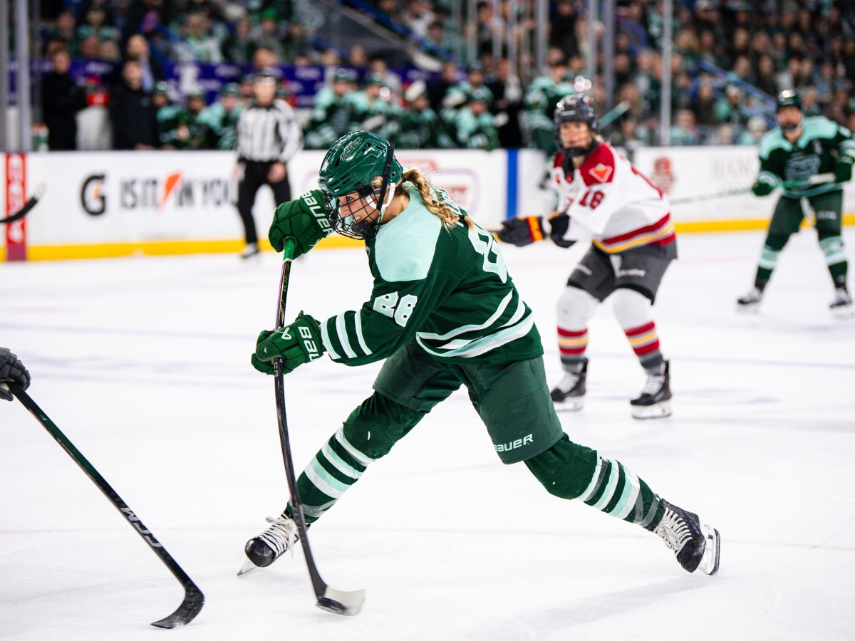 Huber leans forward and flexes her stick as she sends the puck toward the goal. She is wearing a green home uniform.