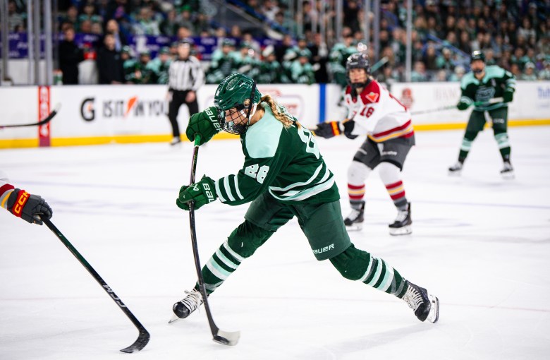 Huber leans forward and flexes her stick as she sends the puck toward the goal. She is wearing a green home uniform.