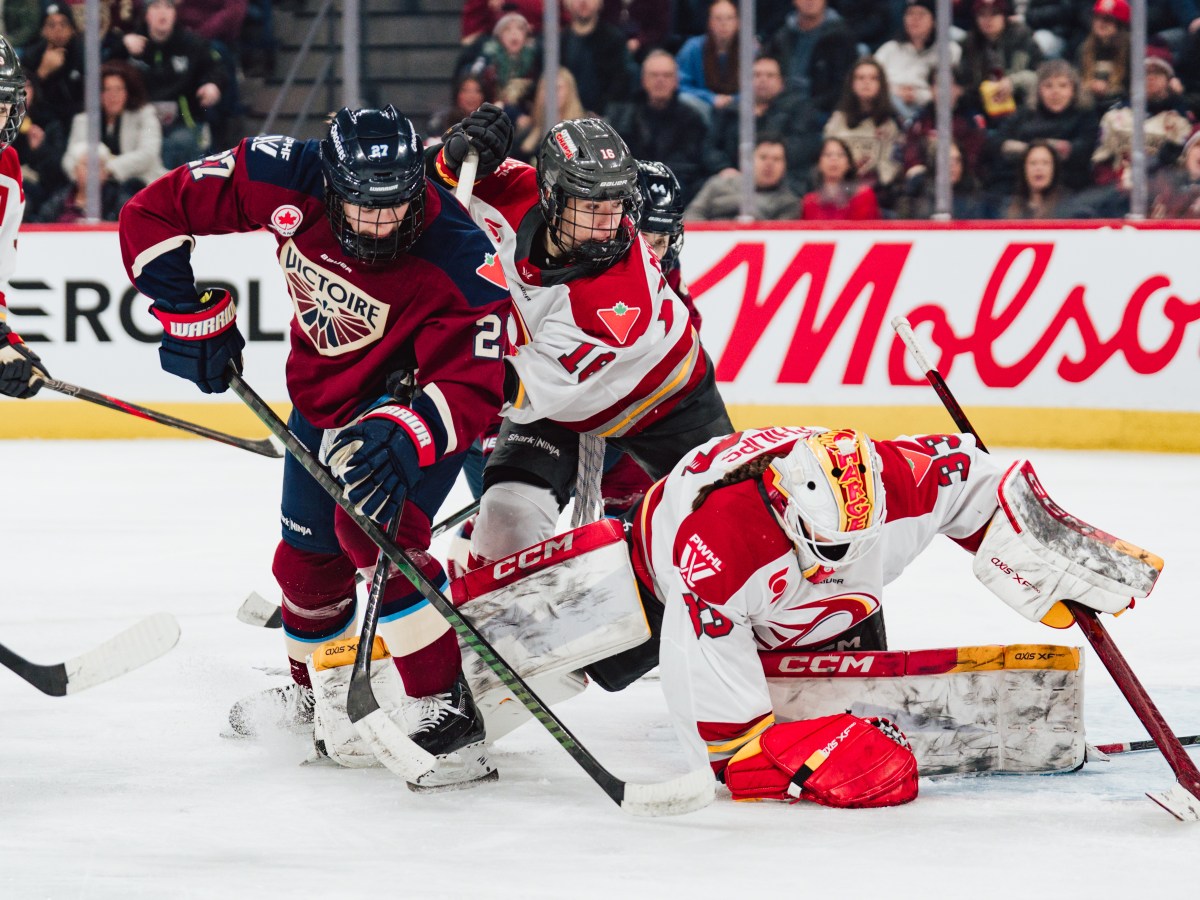 One Montreal Victoire and three Ottowa Charge players scramble for the puck during a game. Fans watch from the stands behind them.