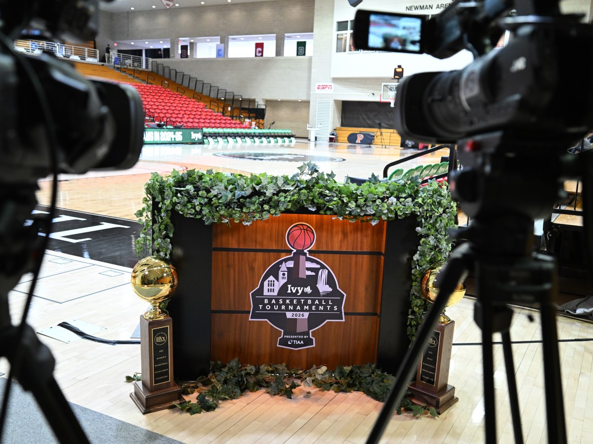 A small podium trimmed with ivy sits near the corner of an empty basketball court. The podium has a large logo on it for the Ivy League basketball tournaments, and two gold trophies are on the floor in front of the podium. Television cameras are visible in the foreground, and the empty court is visible in the background.