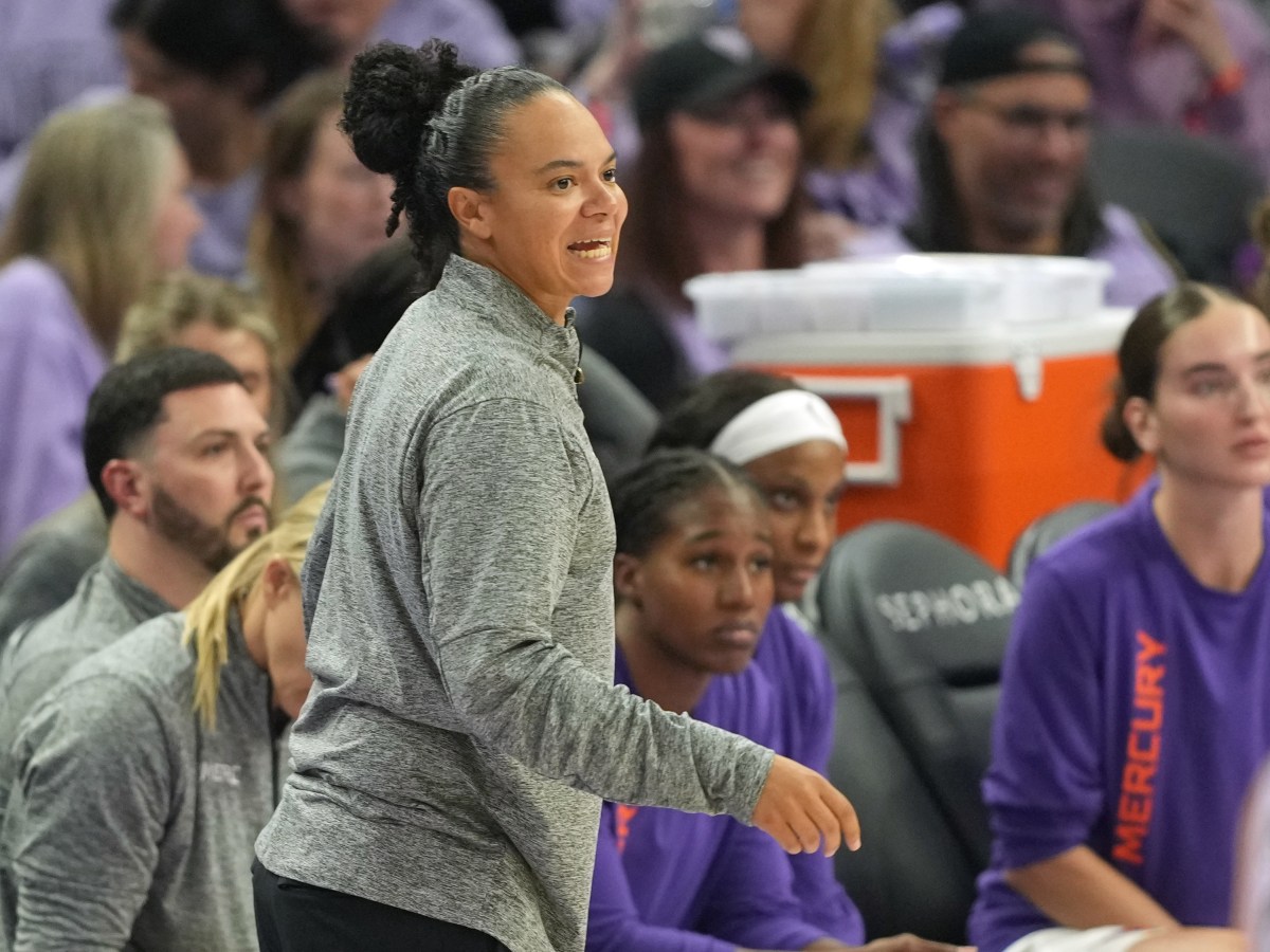 Kristi Toliver, in a gray jacket, enthusiastically guides players on the Phoenix Mercury bench. Players listen intently, and fans watch in the background.