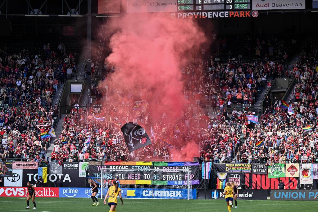 A crowd of Portland Thorns supporters cheer in the stands behind a goal as red smoke rises into the air