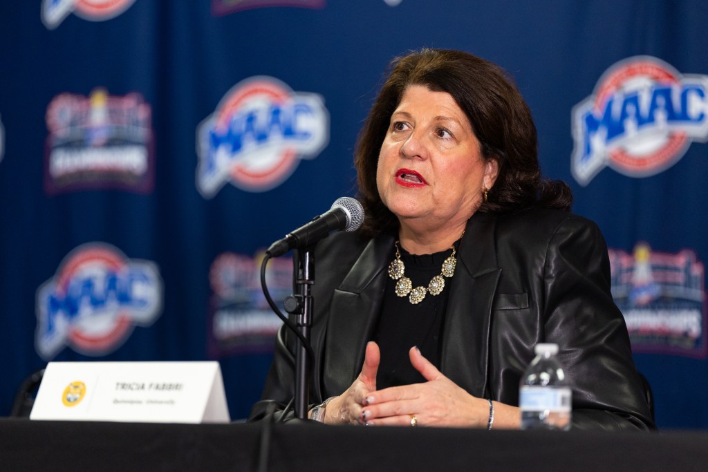 Quinnipiac head coach Tricia Fabbri sits at a podium for a press conference and makes eye contact as she speaks. A microphone and a placard with her name on it are in front of her, and MAAC logos are on a backdrop behind her.