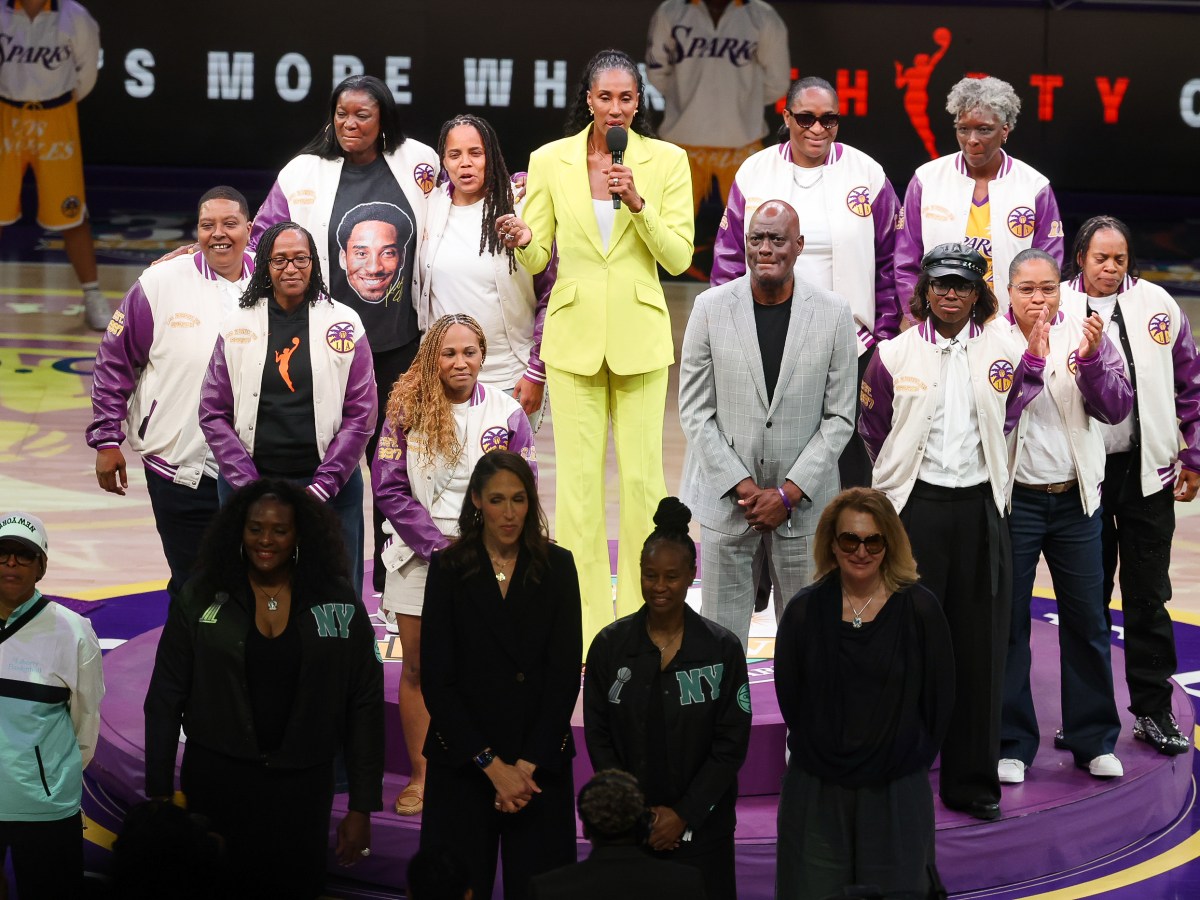 A high angle picture players from the 1997 Los Angeles Sparks team in white letterman jackets with purple Sparks sleeves in a circle around Lisa Leslie who wears a yellow suit and is talking into a microphone.
