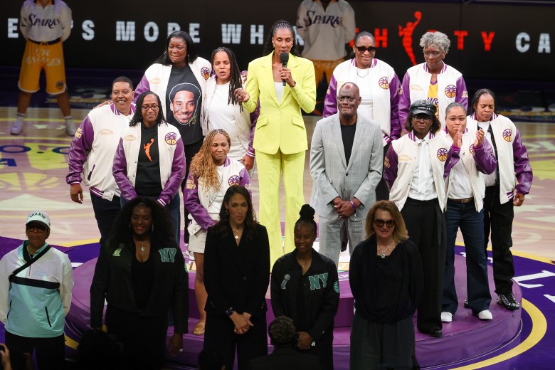 A high angle picture players from the 1997 Los Angeles Sparks team in white letterman jackets with purple Sparks sleeves in a circle around Lisa Leslie who wears a yellow suit and is talking into a microphone.