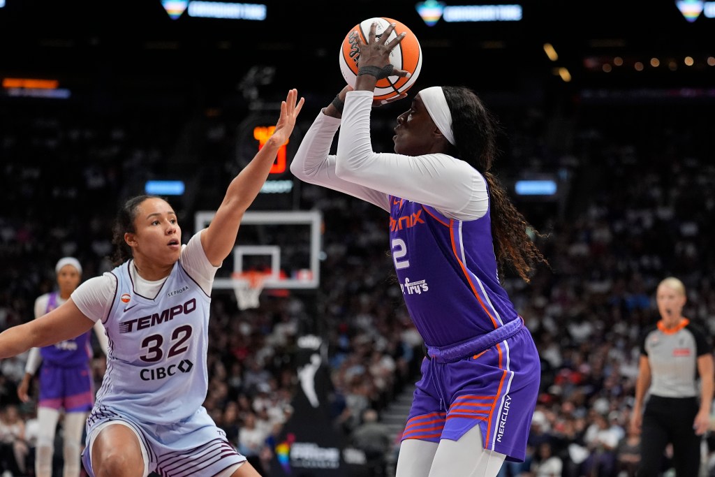Phoenix Mercury wing Kahleah Copper aims the ball over her head to shoot over Toronto Tempo guard Tima Pouve. The crowd watches on in the background. A blurred out Mercury player and referee are also in the background.