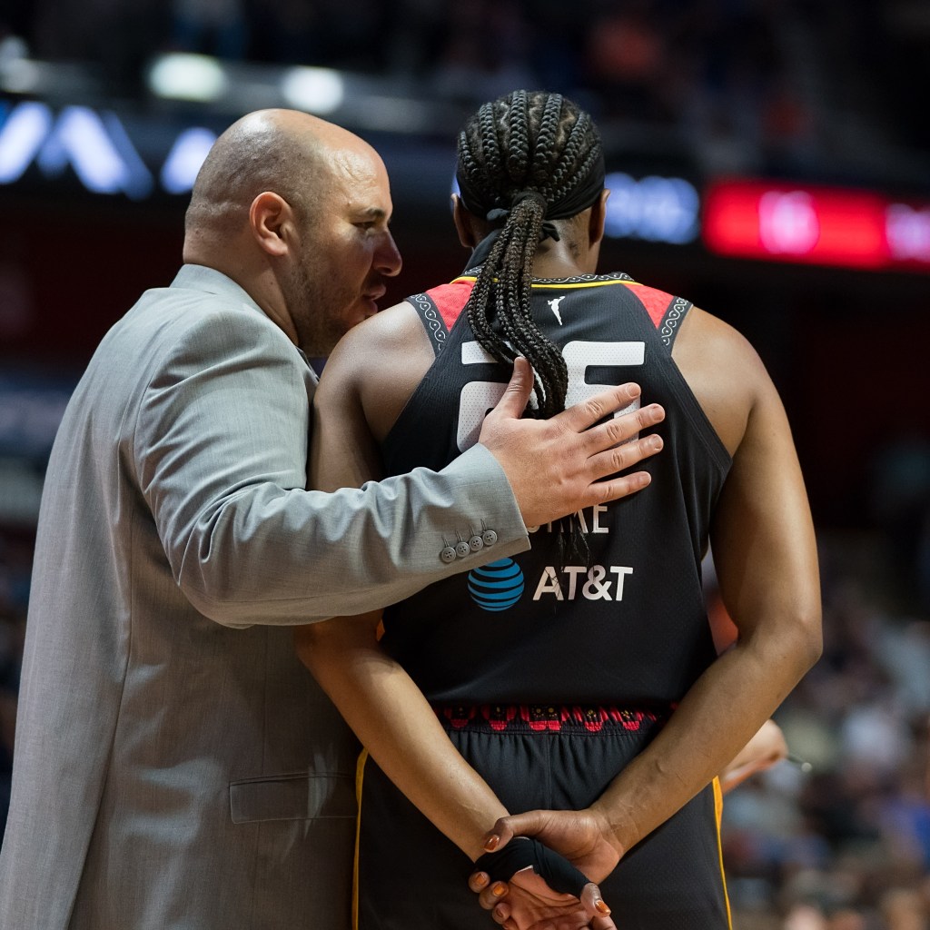Rachid Meziane wearing a grey suit places his hand on the center of Kennedy Burke's back as he talks to her on the sideline. Kennedy Burke also has her hands behind her back.
