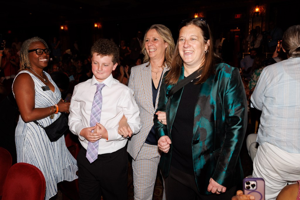 Cheryl Reeve (center) is escorted by her son Oliver (left) and her wife Carley (right) at the Women's Basketball Hall of Fame induction ceremony. They're walking down an aisle and have big smiles on their faces.