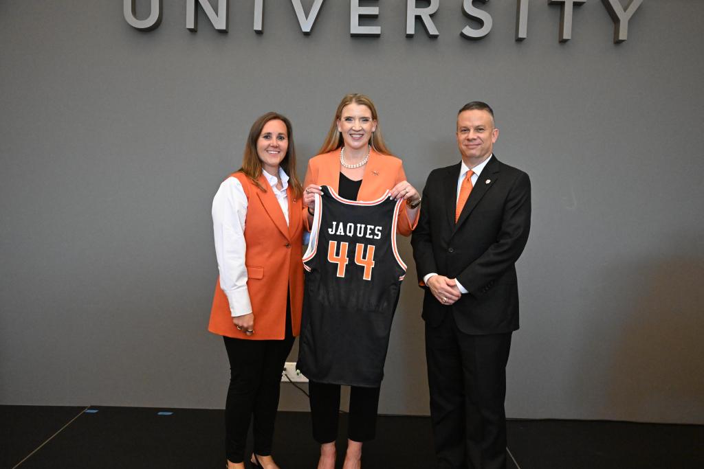 Three people are standing with the lady in the middle holding a black basketball uniform.