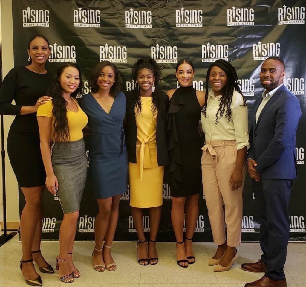 Seven people are smiling while standing in front of a black backdrop adorned with the Rising Media Stars wordmark and logo.