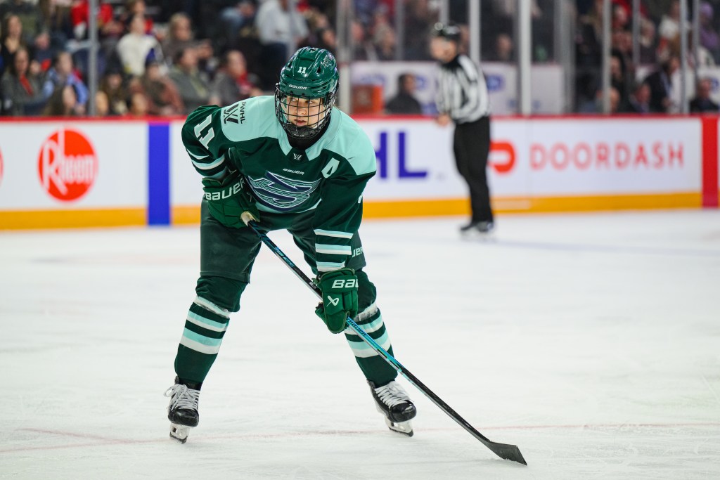 Müller leans on her stick as she lines up on the hashmarks in preparation for a faceoff. She is wearing a green home uniform.