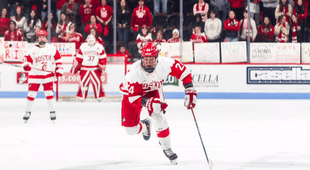 Kelly looks up ice as she skates hard through the neutral zone. She is wearing a white Boston University uniform.