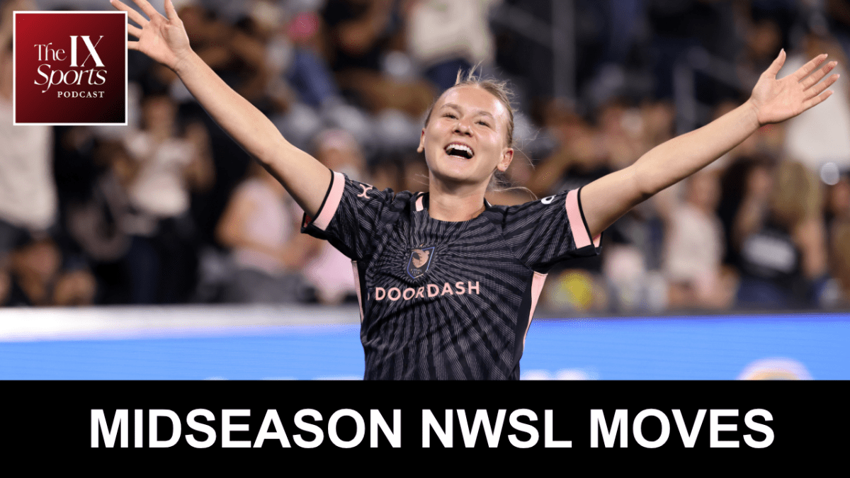 A woman with her hair pulled back and wearing a black soccer kit raises her arms in celebration.