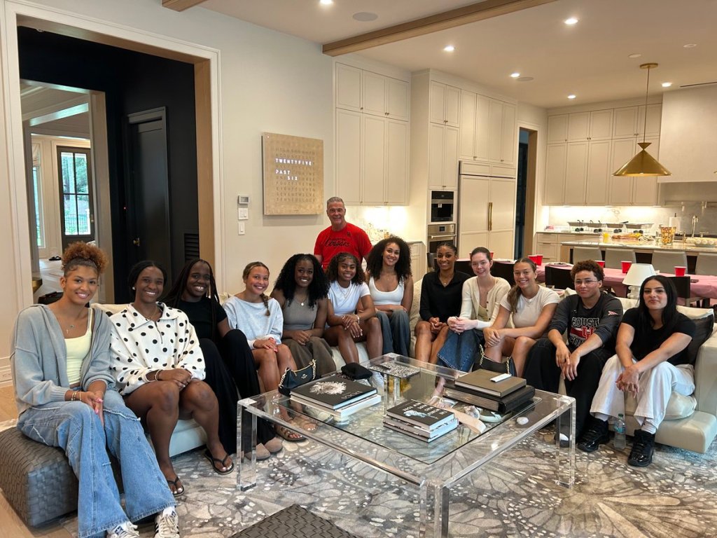 A group of young women sit smiling in a living room with an older male coach standing behind them. 