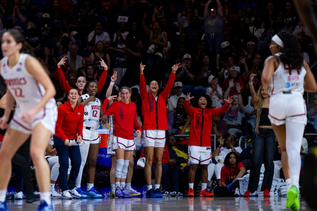 Washington Mystics players celebrate on the bench. Several players hold up three fingers on each hand, and some shout. On the court, guard Sonia Citron gets in a defensive stance, and forward Kiki Iriafen runs down the court.