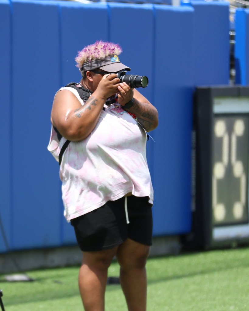 Photographer in white shirt and black shorts with camera up to their face. Wearing a white visor and has purple hair. Standing on a field with a blue backdrop behind them.