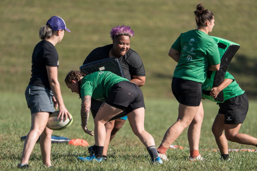Rugby players practicing. Player in black shirt with purple hair holds a tackling pad as someone in black shorts and green shirt tackles the pad and the person.