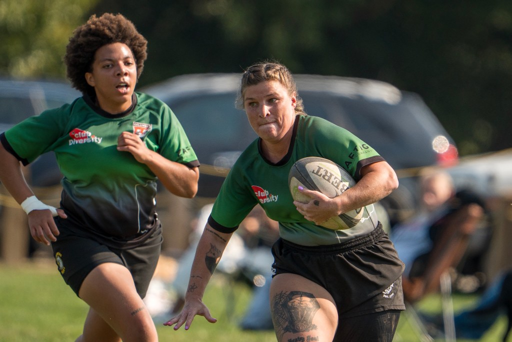 Rugby player holds the rugby ball in her left arm and is running. Has a teammate right behind her. Both wearing green shirts with black shorts.