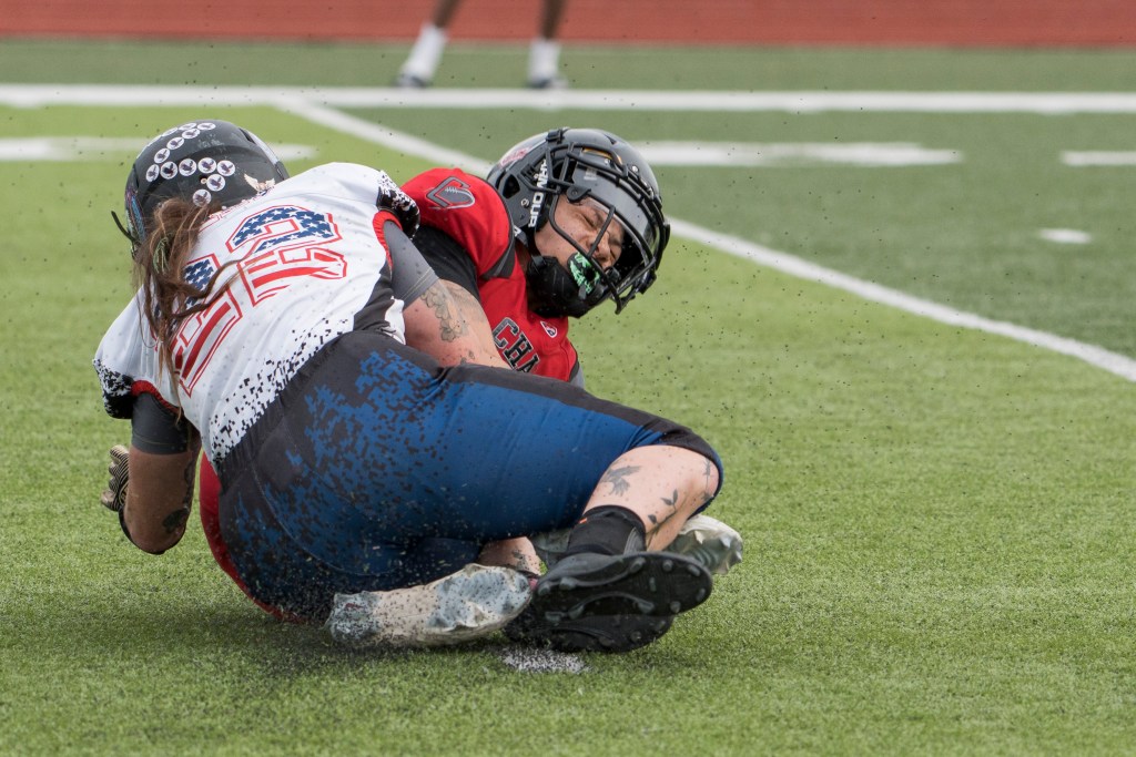 No. 22 in a white jersey and dark blue shorts tackles another football player in red jersey and black helmet. Both players are on the ground.