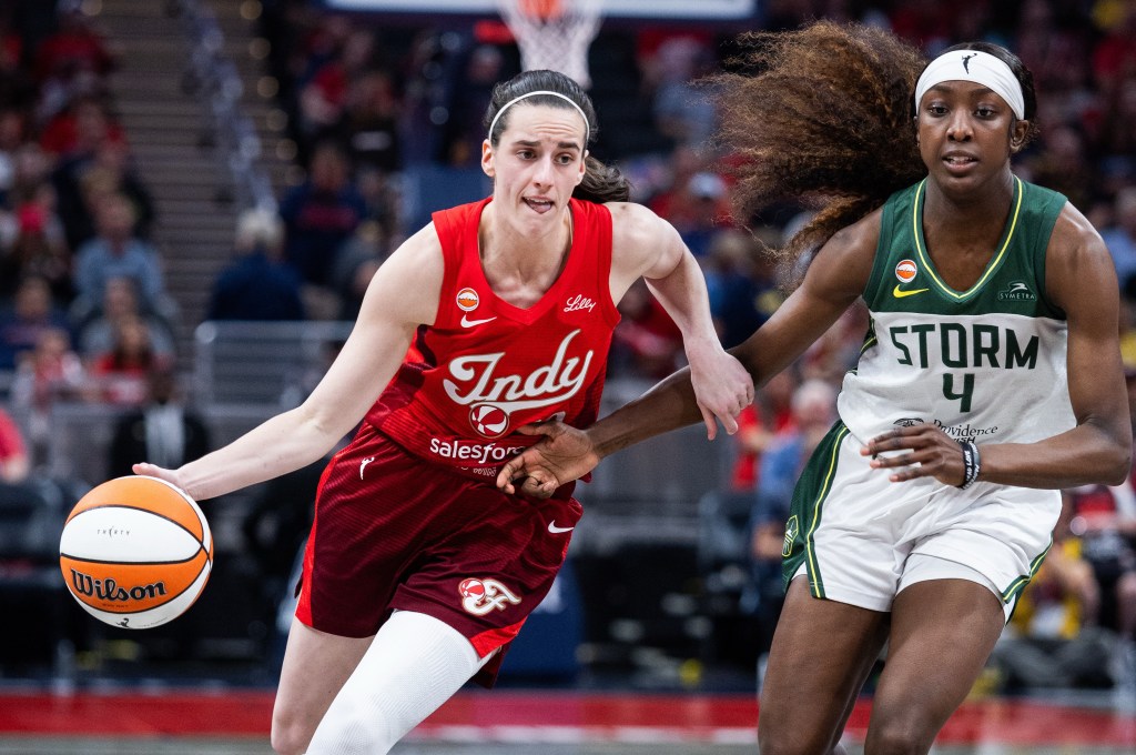 Indiana Fever guard Caitlin Clark dribbles the ball with her right hand. Her tongue is sticking out slightly as she focuses. Seattle Storm guard Flau'jae Johnson tries to stay with Clark, running next to her on her left hip and extending her right arm into Clark's body.