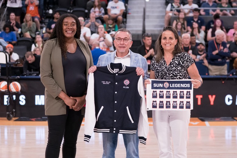 Morgan Tuck wearing a black shirt and pants with an olive green blazer with her hands folded together. She is standing next to Curt Miller who is wearing a light blue suit and his holding up a navy blue and white Connecticut Sun varsity jacket. Jennifer Rizzotti stands to his left wearing white pants and a black and white patterned shirt holding up a miniature white Connecticut Sun Legends banner.