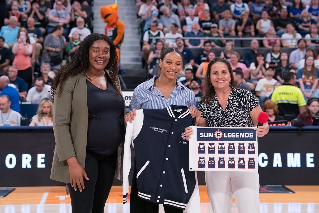 Morgan Tuck wearing a black shirt and pants with an olive green blazer with her hands folded together. She is standing next to Jasmine Thomas who is wearing a light blue shirt and his holding up a navy blue and white-sleeved Connecticut Sun varsity jacket. Jennifer Rizzotti stands to herleft wearing white pants and a black and white patterned shirt holding up a miniature white Connecticut Sun Legends banner. 