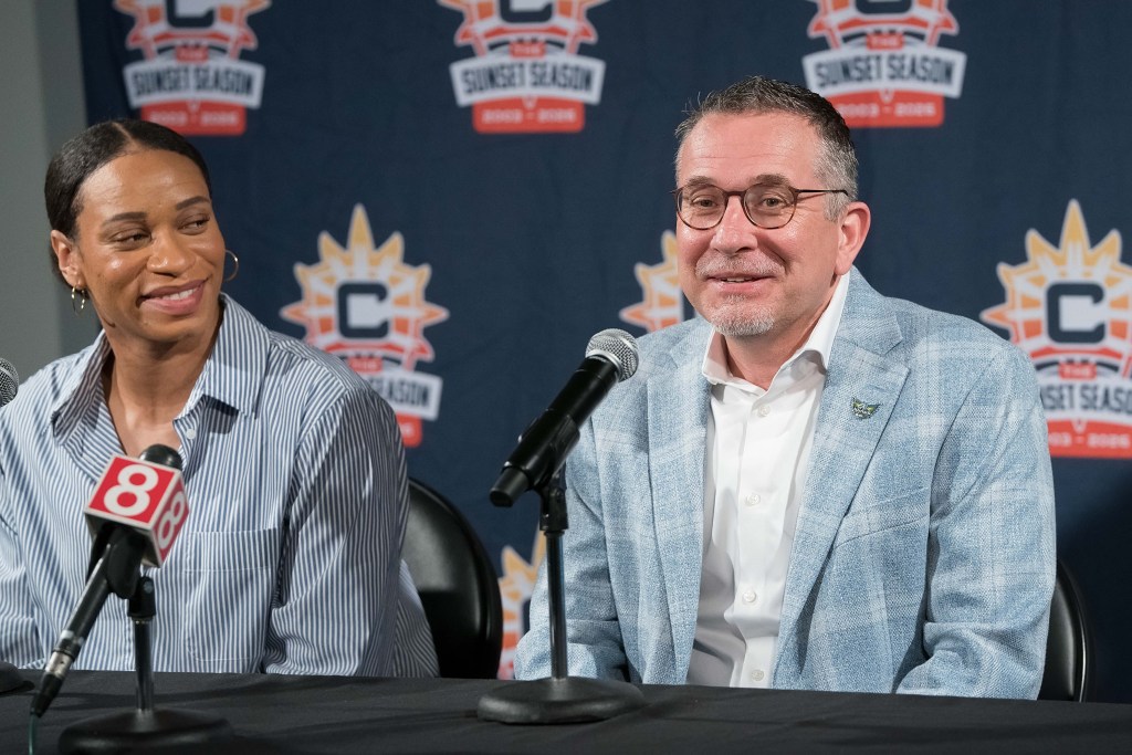 Curt Miller and Jasmine, both in light blue clothing, sit at a table with a black cloth over it in front of  microphones smiling.