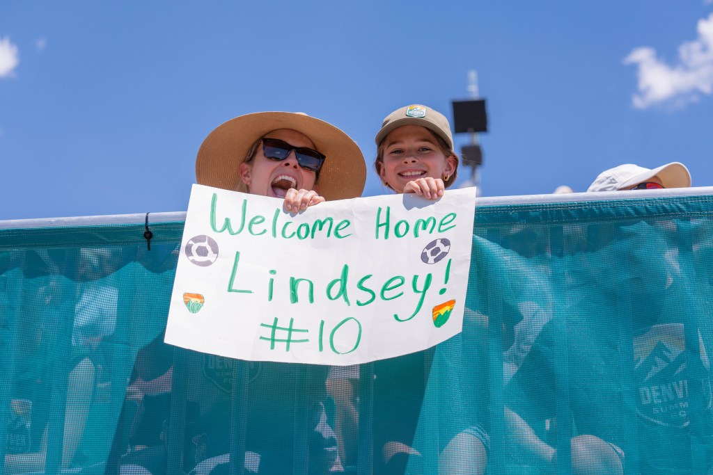 An adult and child smile while holding a sign reading, "Welcome home, Lindsey! #10" with Denver Summit FC logos and soccer balls drawn around the words.