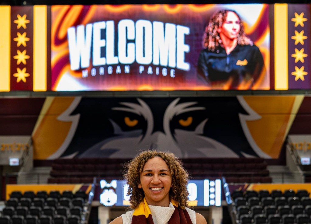 Loyola Chicago head coach Morgan Paige smiles for a photo in front of a giant screen that reads “Welcome Morgan Paige” at Gentile Arena.