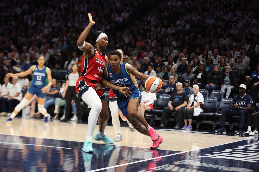 Minnesota Lynx guard Courtney Williams dribbles the ball with her left hand along the baseline. Washington Mystics forward Kiki Iriafen slides her feet as she defends Williams tightly and has her right hand in the air, away from Williams' body.