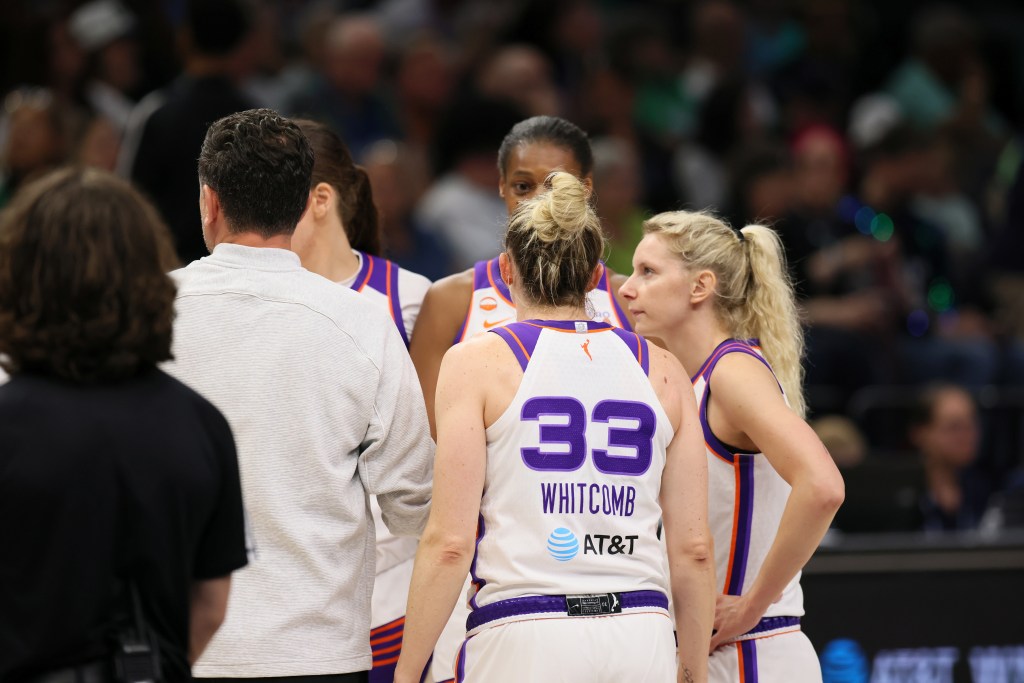 A group of players in white and pink jerseys gather on a basketball court in a team huddle