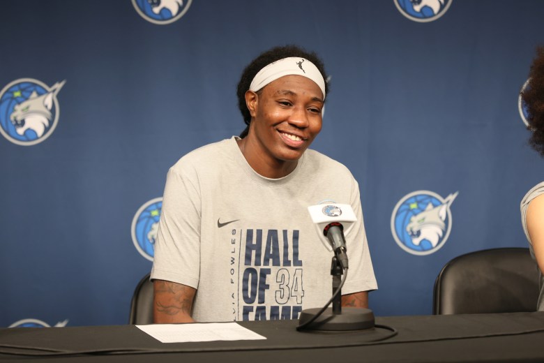 Natasha Howard sits at a table in a gray shirt in front of a blue backdrop with a big smile on her face