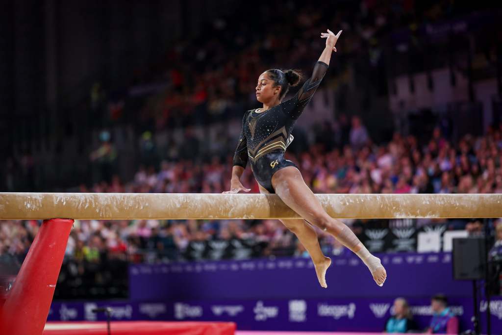 Gymnast Lia Monica Fontaine performs on the balance beam in a black leotard, her legs swinging down and one hand up in the air, in front of an audience at the 2026 Commonwealth Games.