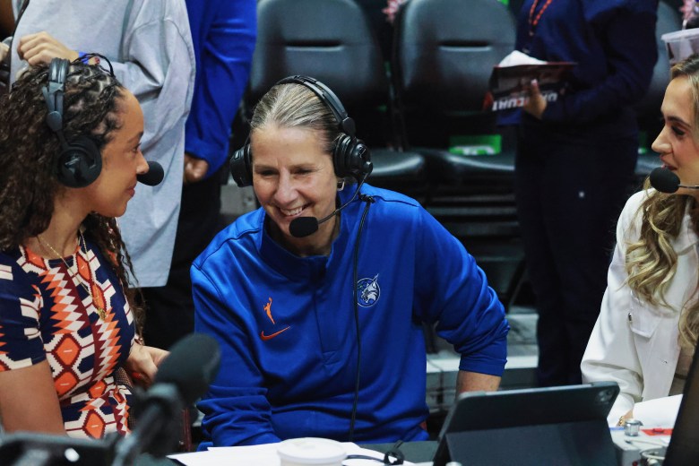 Cheryl Reeve sits between two women in her blue Minnesota Lynx quaterzip wearing a headset with a big smile on her face