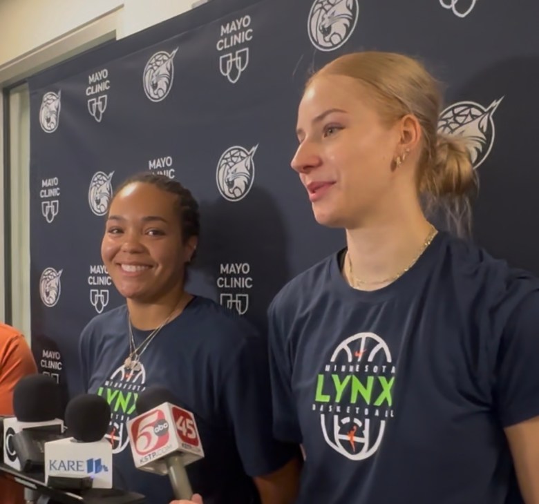 Napheesa Collier and Dorka Juhász stand in front of a dark blue back drop wearing matching dark blue Lynx practice shirts.