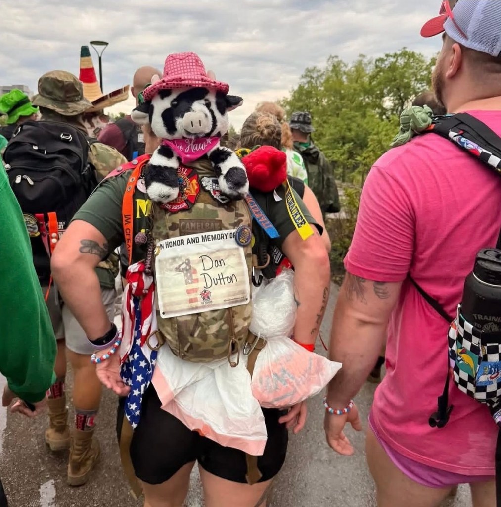 Back of person with backpack on with stuffed animal cow sticking out of the top. Paper on the back of the brown backpack reads "In honor and memory of Dan Dutton"