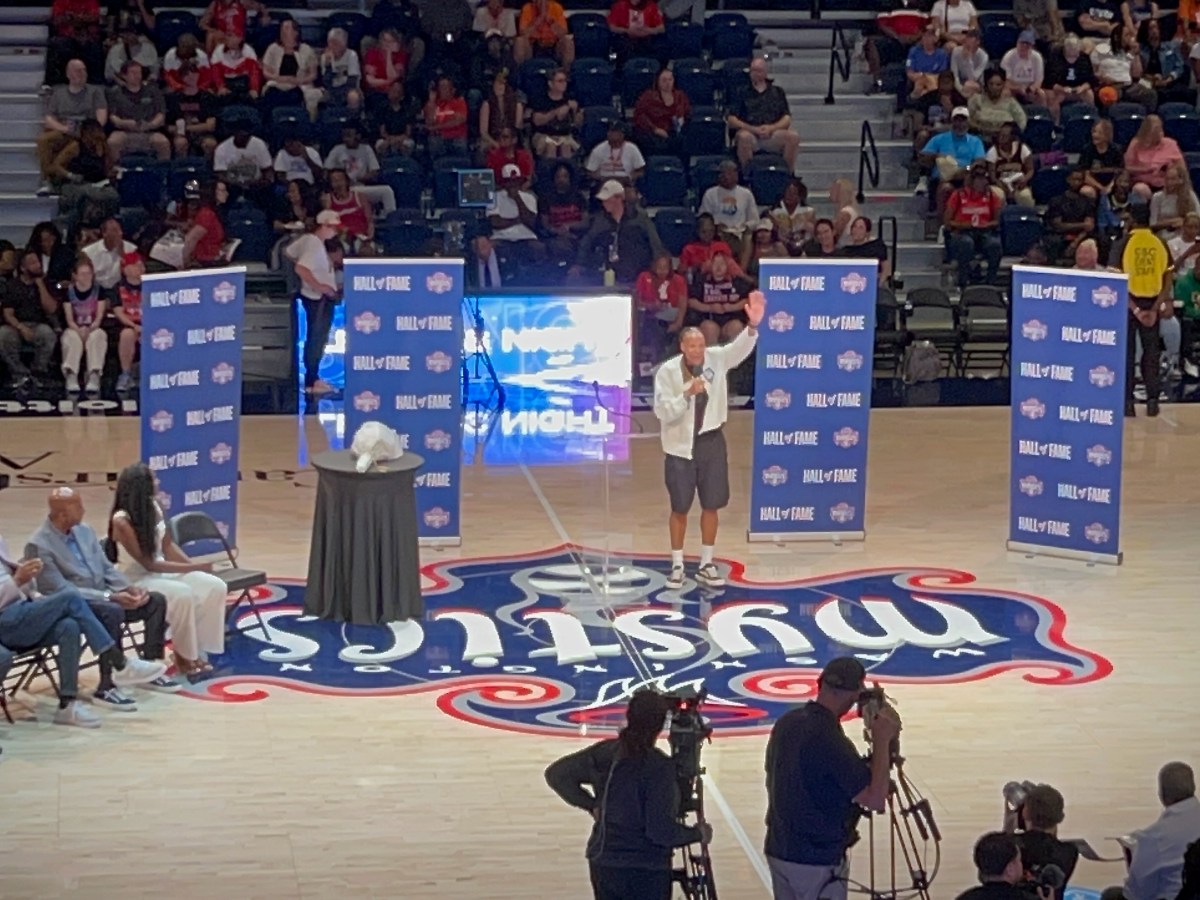 Former Washington Mystics forward Monique Currie holds a microphone in one hand and waves to the crowd with the other. She is standing at center court. Behind her are several dark blue backdrops with the Mystics logo and "Hall of Fame" repeating on them. Her family and other people participating in her Hall of Fame induction ceremony are seated to her right.