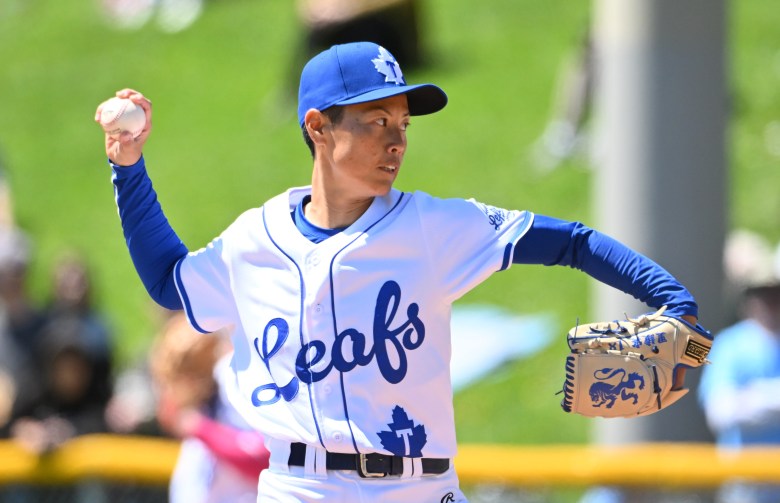 A woman in a white baseball jersey with blue script that says "Leafs" winds up a pitch with her right hand.