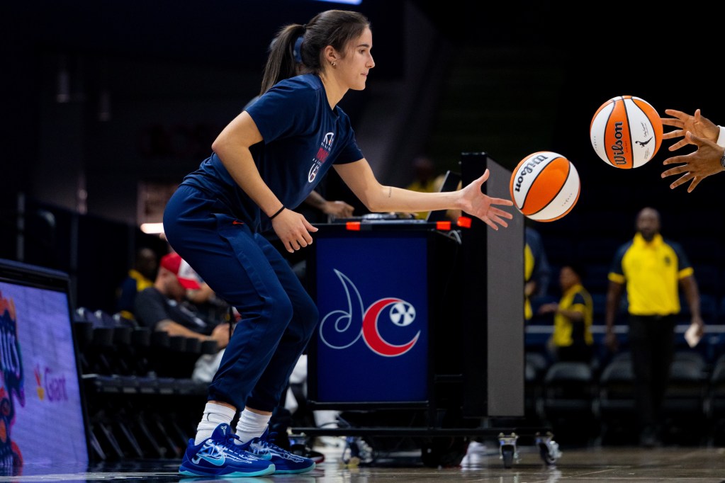 Washington Mystics guard Sonia Citron is shown from the side, doing a ball-handling drill where she has to pass one basketball while catching another. She is wearing a navy T-shirt and navy sweatpants.