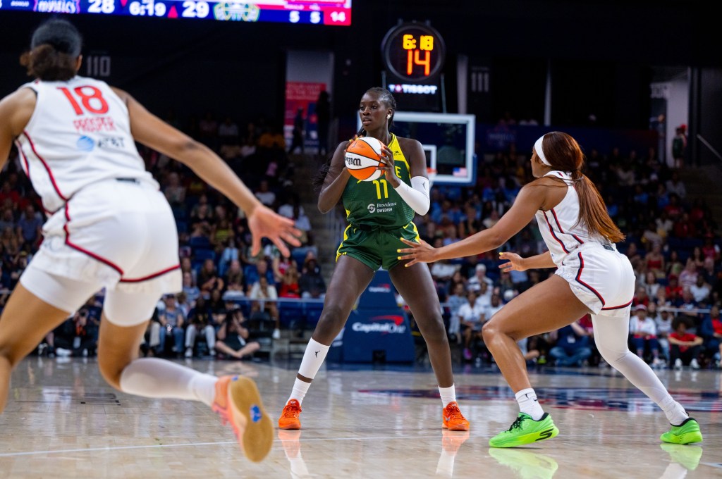 Seattle Storm center Awa Fam holds the ball behind the 3-point line. Washington Mystics forward Kiki Iriafen is in a defensive stance guarding her, while guard Cassandre Prosper is ready to help if needed.