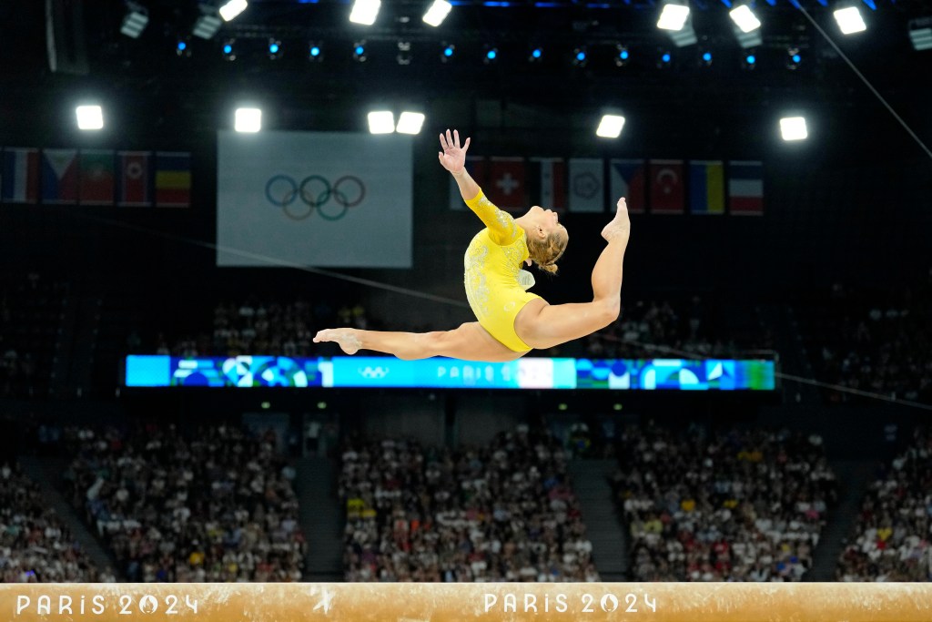 Flavia Saraiva, wearing a gold leotard, jumps with her head back above the balance beam in front of an Olympic flag
